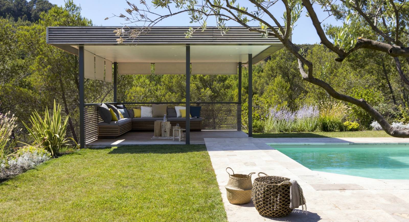 Image of a pergola shading an outdoor sitting area in the corner of a lush garden nearby an in-ground swimming pool.