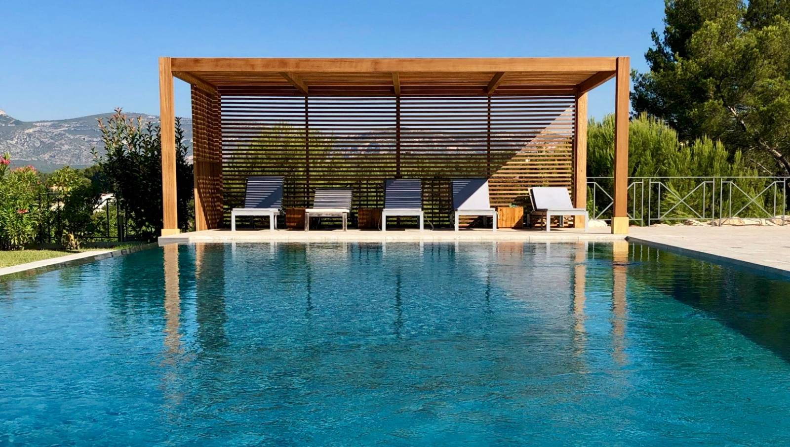 Image of a in-ground swimming pool with covered lounge chairs and a wood slat pergola shading the patio.