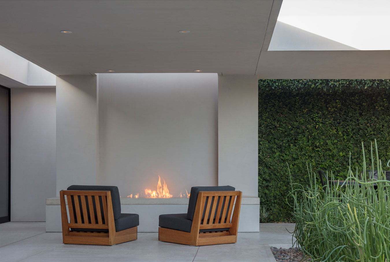Defined by a smooth light-colored flooring that contrasts with a rich green vertical garden, wooden armchairs invite relaxation and conversation, positioned to face the warmth of the elongated, minimalist fireplace embedded in a clean, white wall.