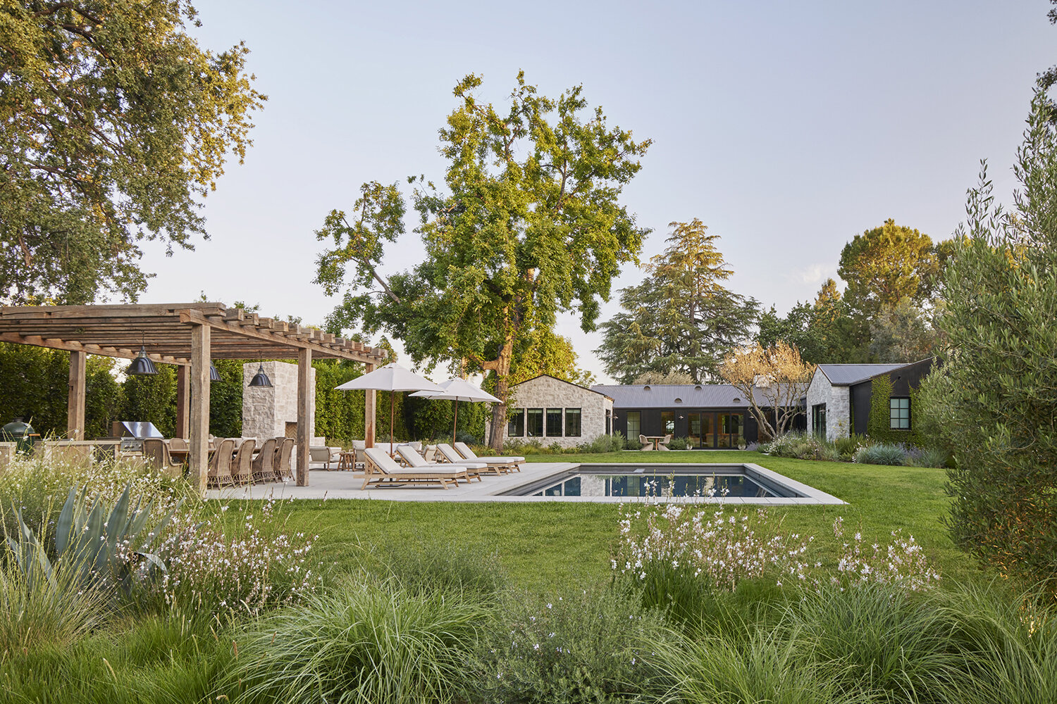 GSLA Studio creates lush backyard with open lawn spaces around a in-ground swimming pool adjacent to a outdoor kitchen area with fireplace and seating.