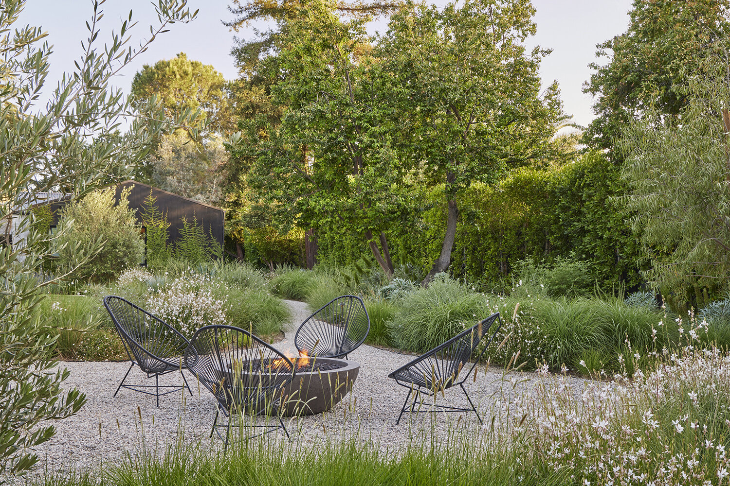 GSLA Studio surrounds firepit and wicker chairs with meandering crushed gravel paths and finely textures native plants consisting of ornamental grasses and wildflowers.