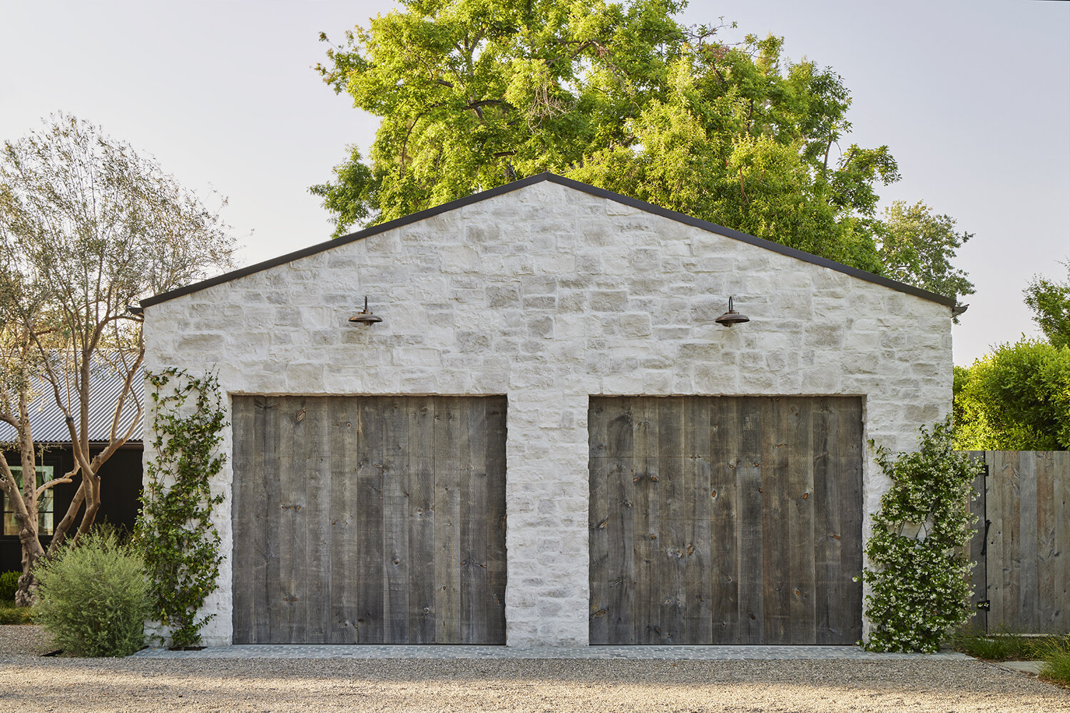 Amber Lewis uses natural materials like white washed stone and reclaimed grey weathered wood for garage doors while GSLA creates naturalistic gardens around the sides.